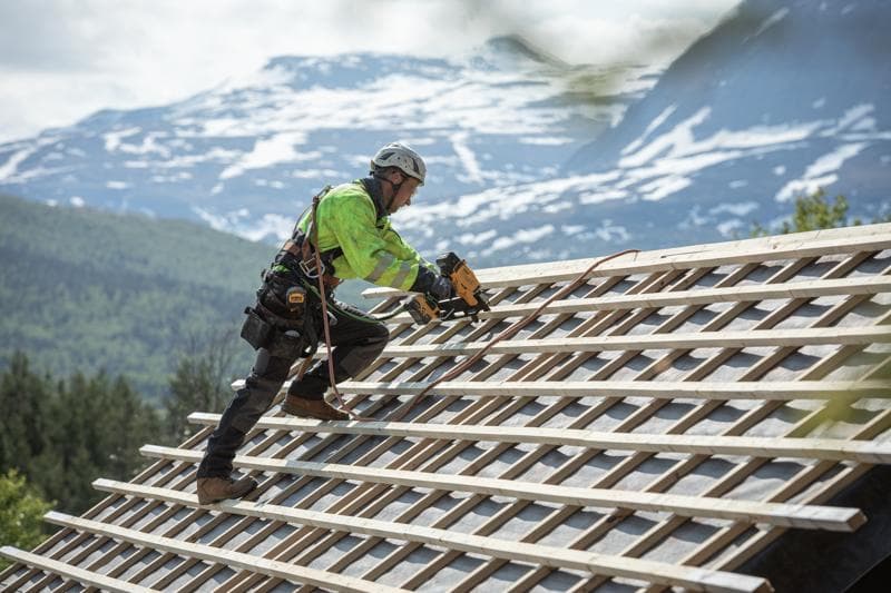 Trabajador usando clavadora DEWALT en techo inclinado al aire libre, con equipo de seguridad y montañas detrás.