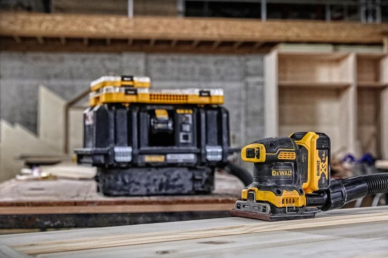 Lijadora inalámbrica DEWALT sobre madera, caja de almacenamiento al fondo en taller.