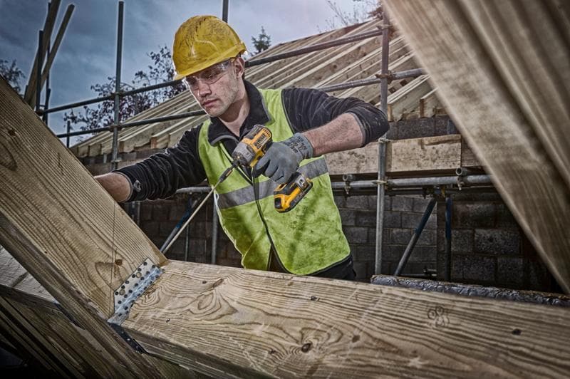 Un trabajador de la construcción con casco amarillo y chaleco reflectante utiliza un taladro inalámbrico DEWALT para fijar
