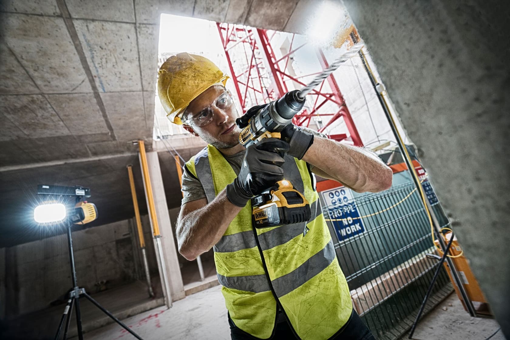 Un trabajador de la construcción con chaleco de seguridad amarillo, guantes y casco utiliza un taladro inalámbrico DEWALT (modelo DCD996P2) en una obra. Se ven luces industriales y materiales de construcción al fondo.