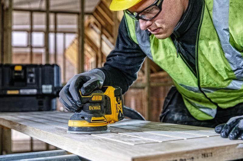 DEWALT DCW210, DCW210D1 lijadora en uso sobre madera, obra, persona con guantes y chaleco de seguridad.