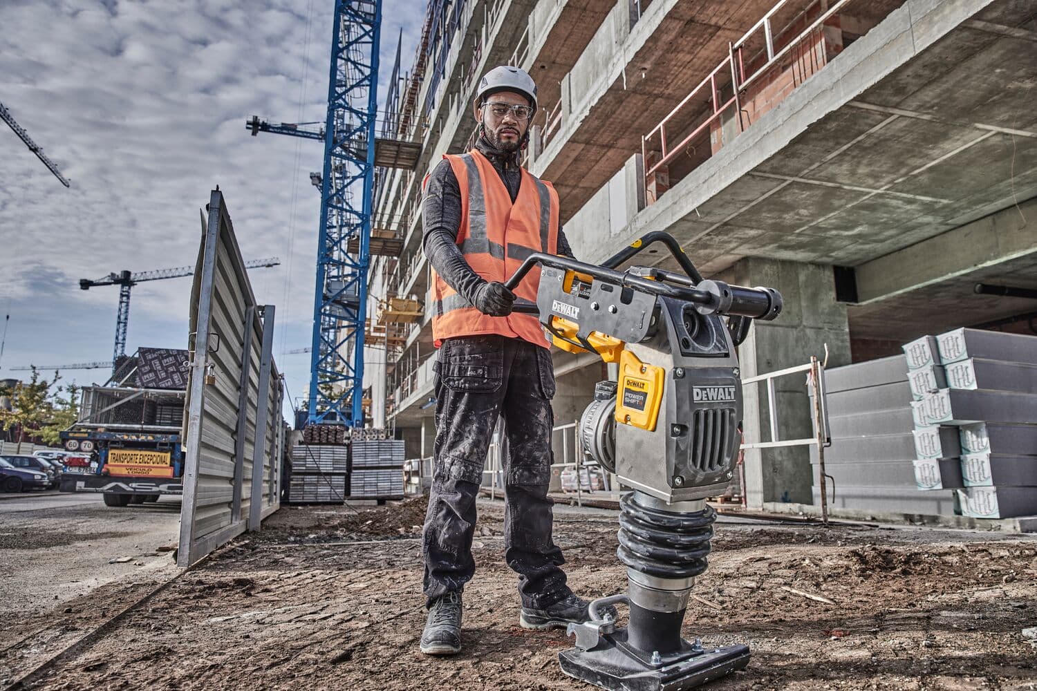 Persona con equipo de seguridad usa un compactador DEWALT en obra, con grúas y edificio en el fondo.