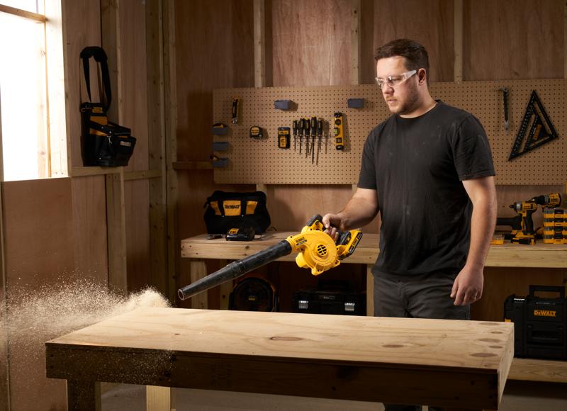 Persona usa soplador DEWALT DCV100 para limpiar aserrín de un banco de madera en un taller.