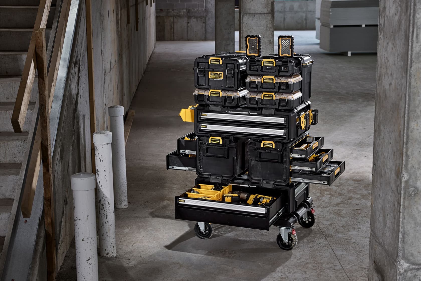 Un carro de almacenamiento de herramientas móvil DEWALT con varias cajas de herramientas negras y amarillas apiladas, cajones y estuches, lleno de diferentes herramientas, situado en un espacio industrial de concreto.