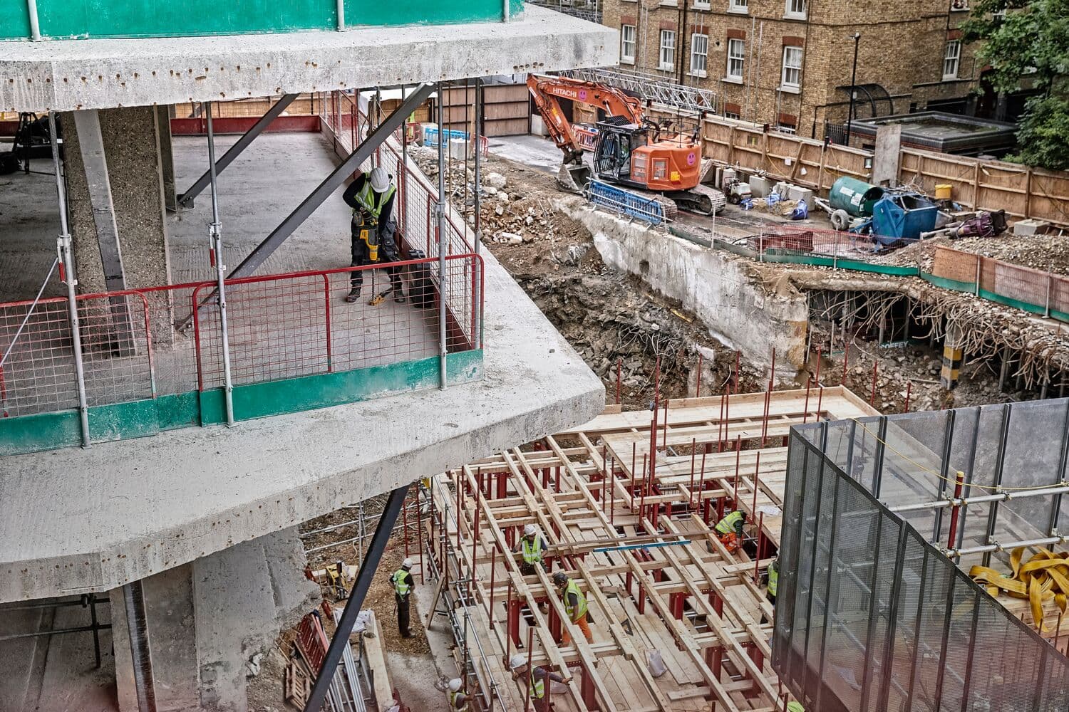 Obra con trabajadores, estructuras de concreto, andamios y excavadora en una zona urbana.
