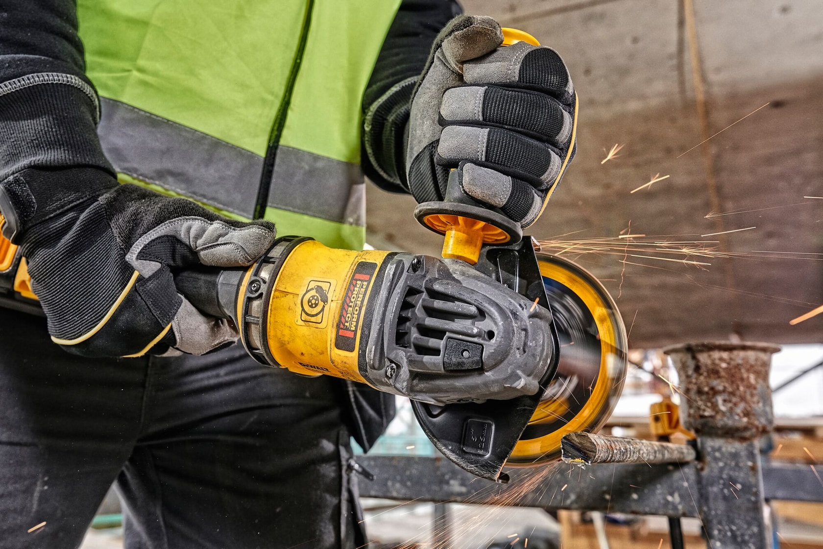 Trabajador usa amoladora DEWALT DCG418X1 en metal, con guantes y chaleco, saltando chispas.