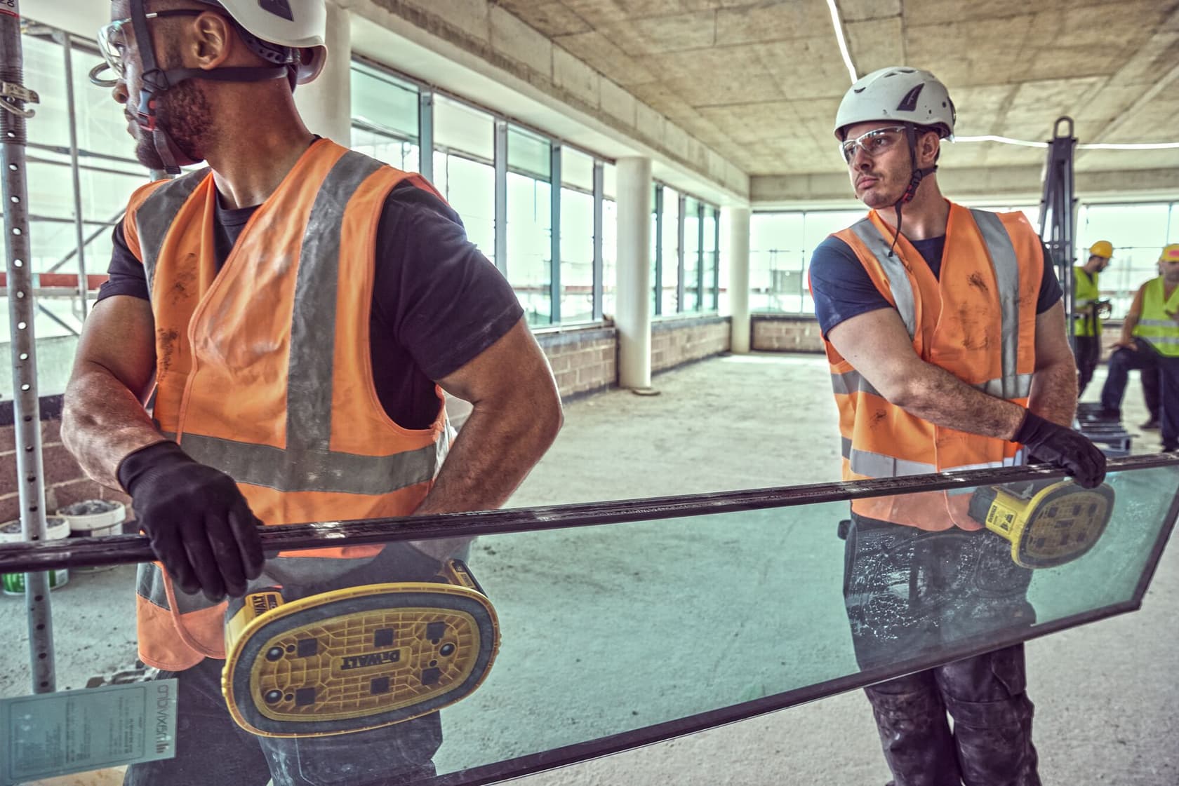 Dos trabajadores mueven vidrio con herramientas DEWALT, usando equipo de seguridad y chalecos naranjas.